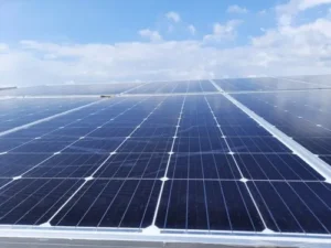 A close-up, low-angle shot of several dark blue monocrystalline solar panels installed on a flat roof under a bright, slightly cloudy blue sky.