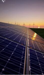 A close-up, angled view of blue solar panels in a field with wind turbines in the background during a vibrant sunrise over the ocean.